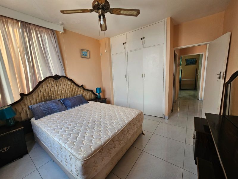 A bedroom featuring tiled flooring, built-in cupboards, and a ceiling fan, offering a functional space with potential for modern updates.