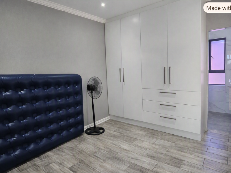 A modern bedroom featuring sleek white built-in cupboards, grey tiled flooring, and recessed ceiling lighting, with an adjoining en-suite bathroom visible.