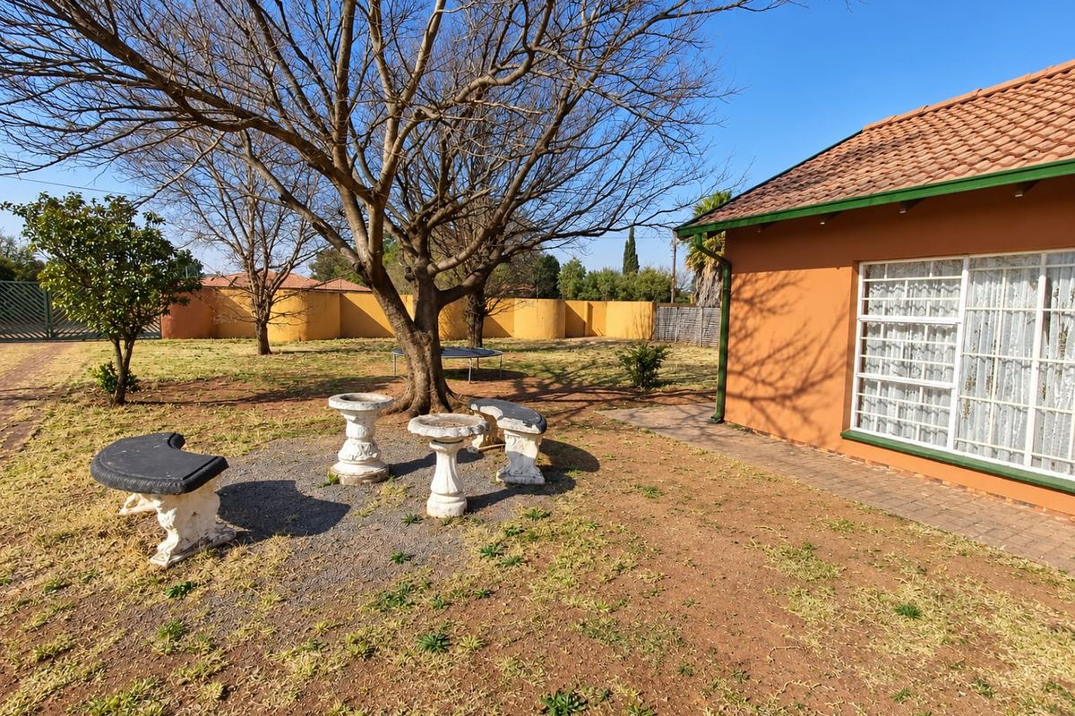 A view of the rear garden area featuring a paved walkway, mature trees, and the exterior wall of the house with a tiled roof.