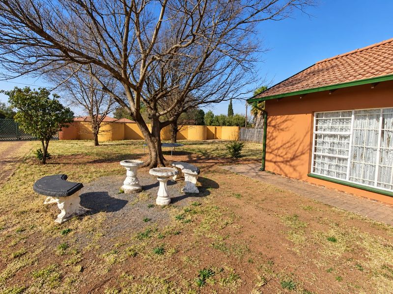 A view of the rear garden area featuring a paved walkway, mature trees, and the exterior wall of the house with a tiled roof.