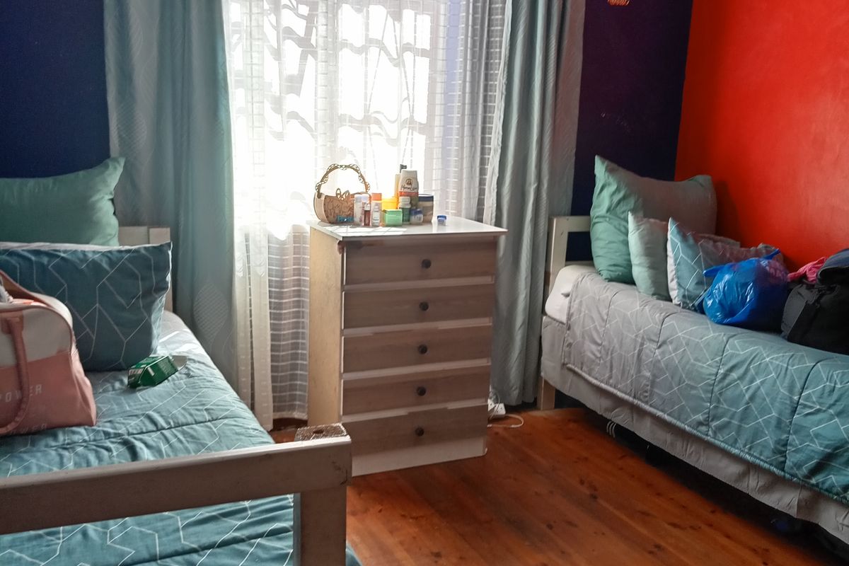 A bedroom featuring timber flooring and a ventilation grille, currently configured with two beds and a central chest of drawers.