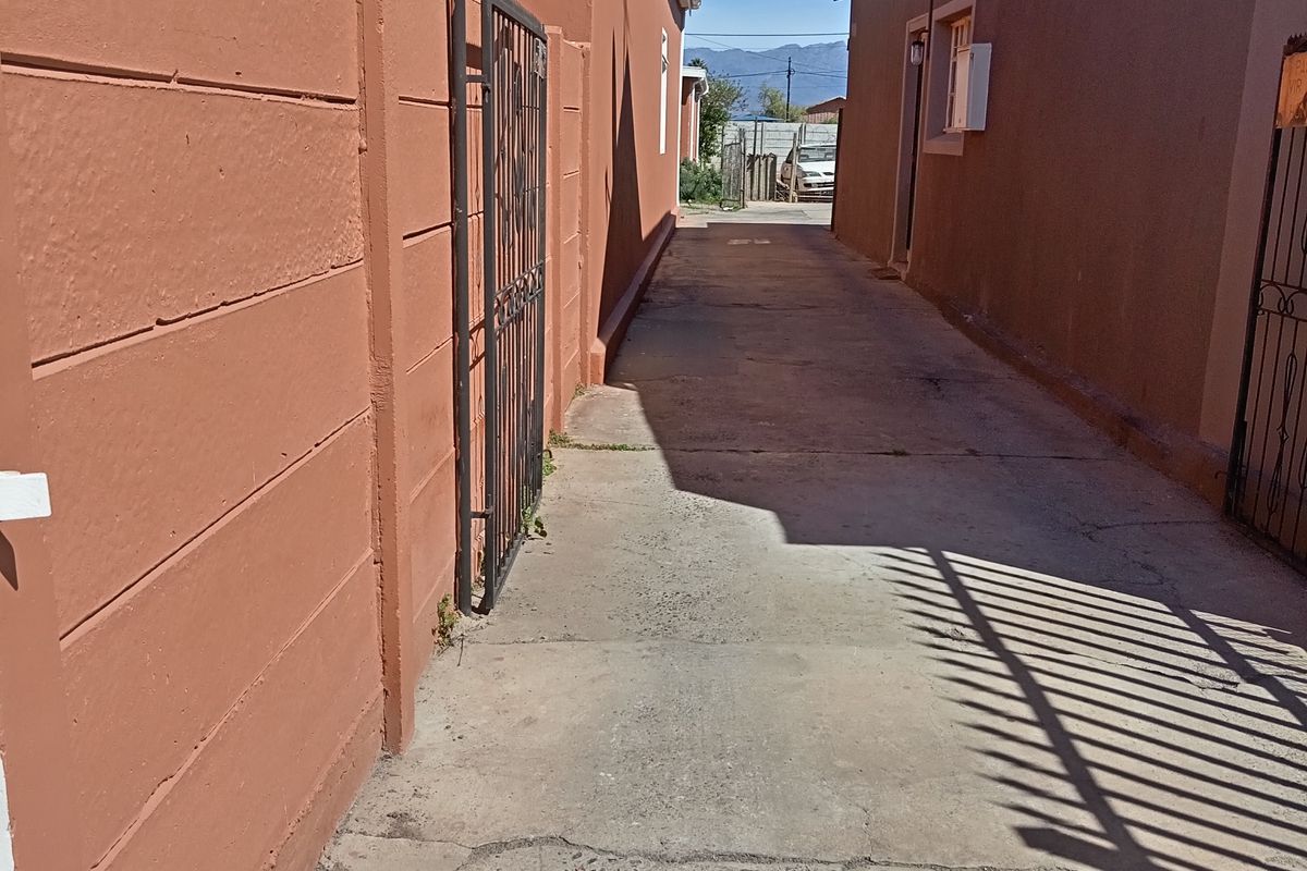 A narrow concrete side passage or driveway area between two residential buildings, featuring boundary walls and metal security gates.