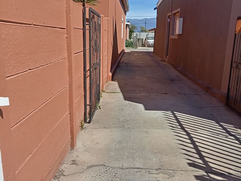 A narrow concrete side passage or driveway area between two residential buildings, featuring boundary walls and metal security gates.