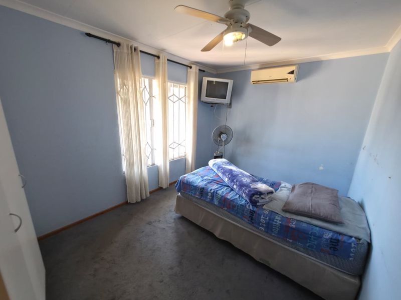 A bedroom featuring a ceiling fan, wall-mounted air conditioner, and security bars on the windows. The room includes built-in storage and carpeted flooring.