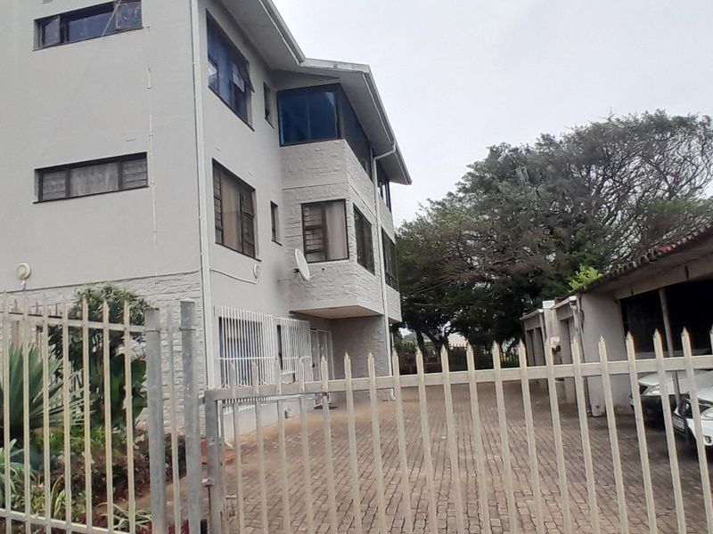 Exterior view of a multi-story apartment building featuring a paved driveway, covered carport parking, and perimeter security fencing.