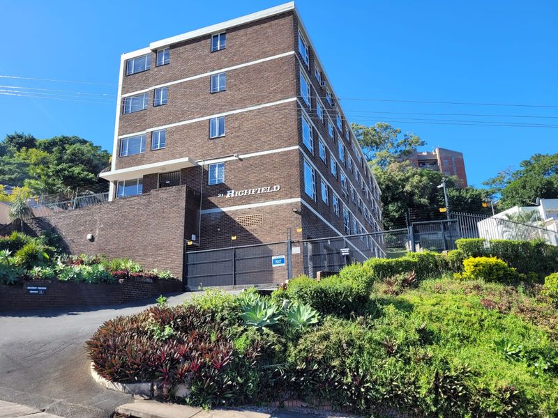 Exterior view of the Highfield apartment building featuring a brick facade, secure gated entrance, and well-maintained landscaped garden area.