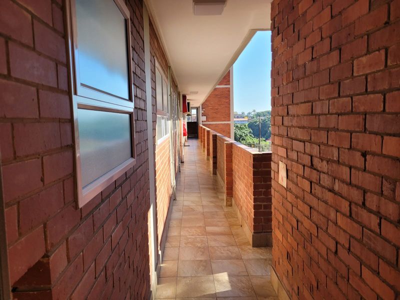 An open-air apartment hallway featuring exposed brick walls, tiled flooring, and a brick balustrade overlooking the surrounding area.