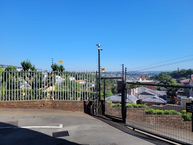 A secure entrance area featuring a metal gate, electric fencing, security camera, and a brick retaining wall overlooking the surrounding neighborhood.