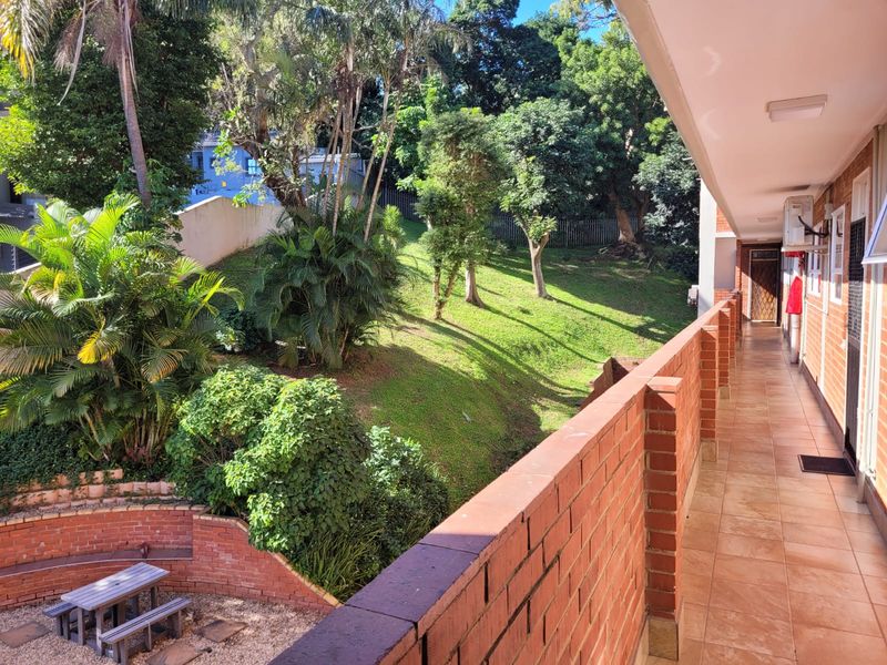 A spacious covered balcony featuring a brick balustrade and tiled flooring, overlooking a lush, landscaped garden area.