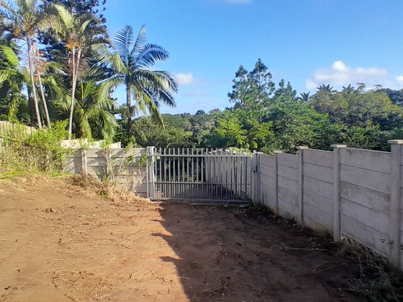 A vacant residential plot featuring a precast concrete boundary wall, timber fencing, and a metal security gate, surrounded by established trees and vegetation.