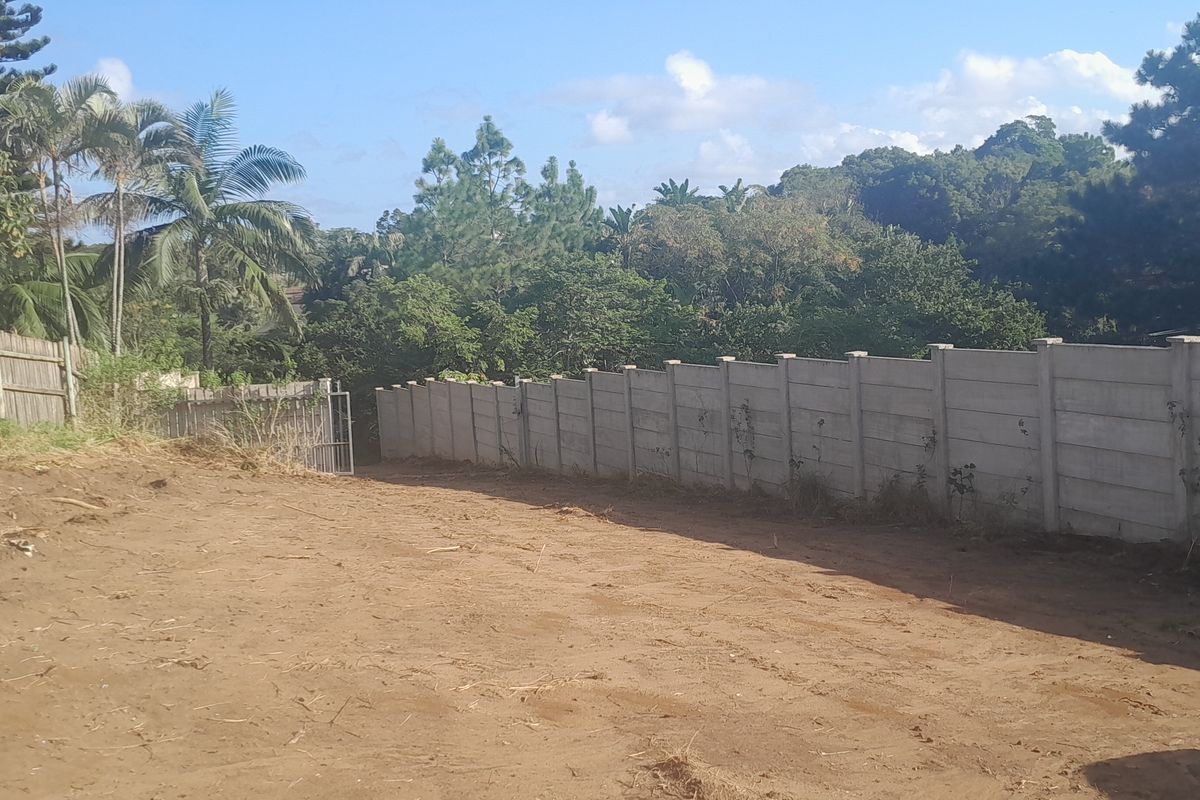 A vacant residential plot featuring a precast concrete boundary wall, timber fencing, and established surrounding greenery.