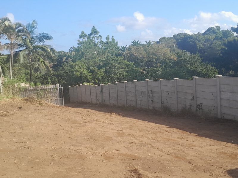 A vacant residential plot featuring a precast concrete boundary wall, timber fencing, and established surrounding greenery.
