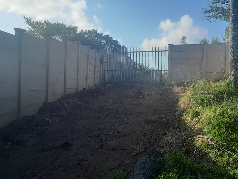 A view of the property entrance featuring a precast concrete boundary wall, a metal security gate, and an unpaved driveway area.