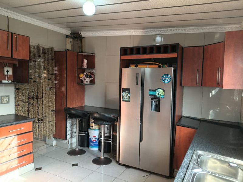 A dated kitchen featuring wood-tone cabinetry, dark laminate countertops, a double stainless steel sink, and tiled flooring with a tiled backsplash.