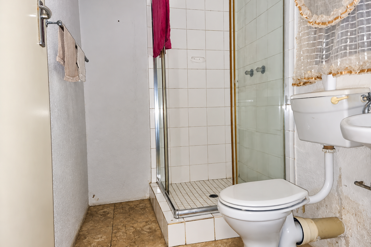 A dated bathroom featuring a shower enclosure, toilet, and washbasin, showing signs of wear and water damage on the lower wall sections.