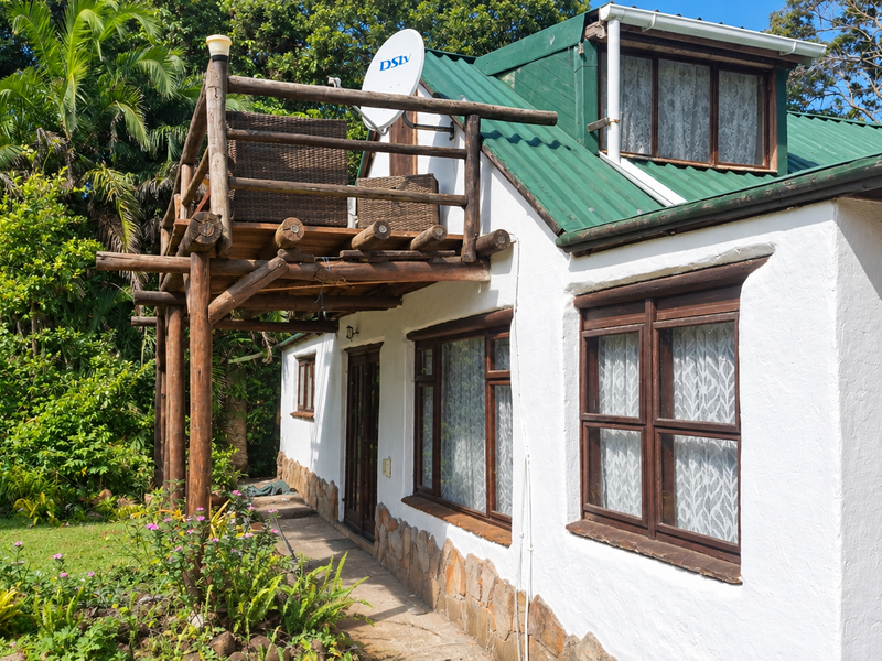 Rear exterior view of the house featuring a timber balcony, corrugated metal roof, and stone cladding accents set against a lush garden.