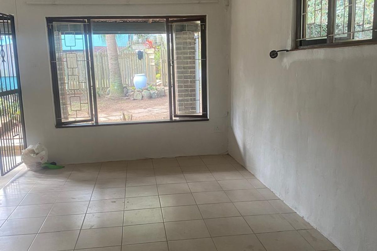 An empty, neutrally painted living room featuring tiled flooring, security bars on the windows, and a sliding door leading to the garden.