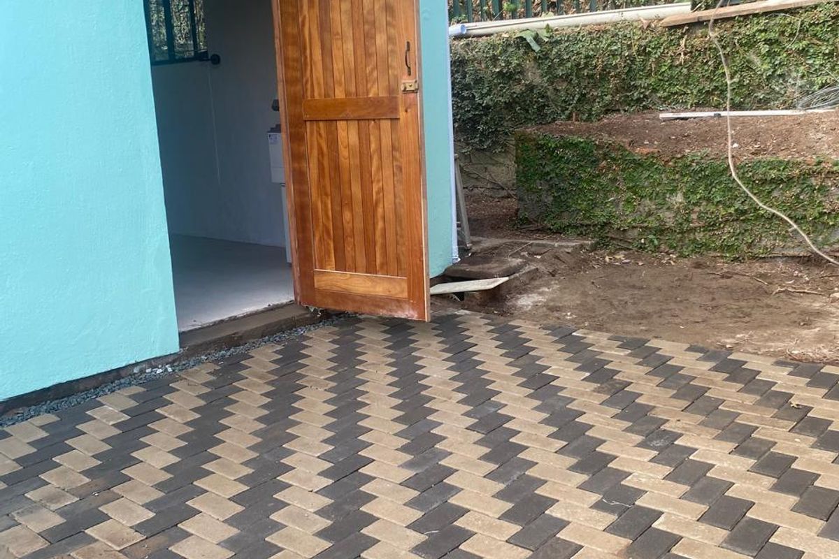 A rear exterior view of a cottage featuring a newly laid herringbone paved patio, a timber entrance door, and a protective polycarbonate awning.