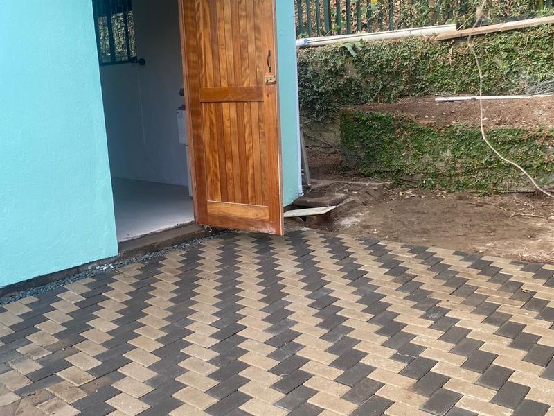 A rear exterior view of a cottage featuring a newly laid herringbone paved patio, a timber entrance door, and a protective polycarbonate awning.
