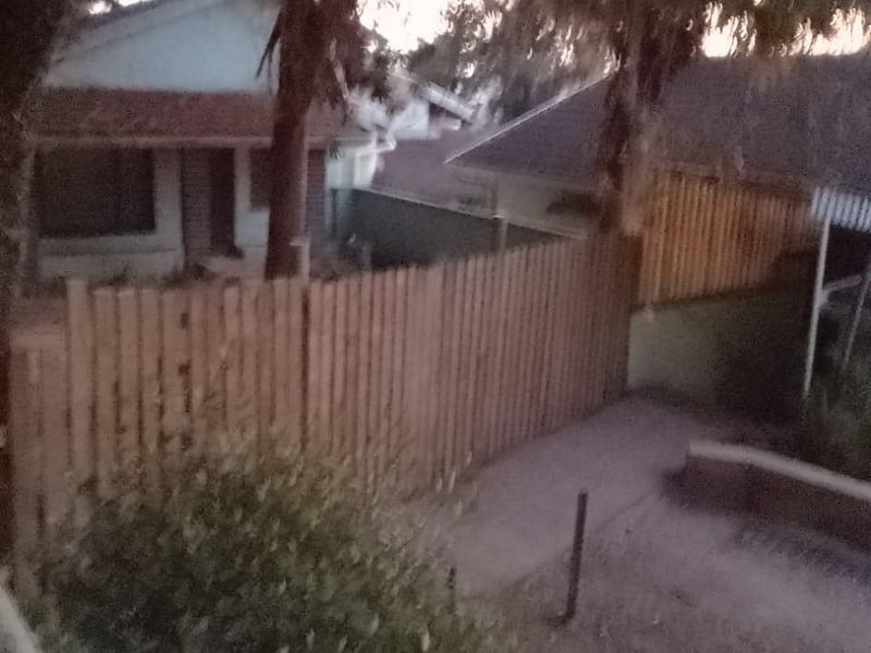 A rear exterior view of the cottage property featuring a timber fence, concrete pathway, and tiled roof structure at dusk.