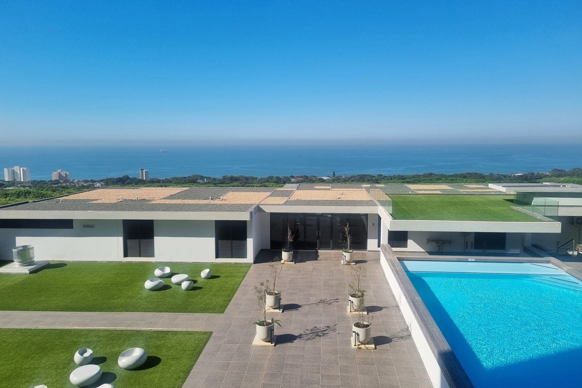 Aerial view of a modern apartment complex featuring a swimming pool, landscaped artificial lawn, and expansive ocean views.