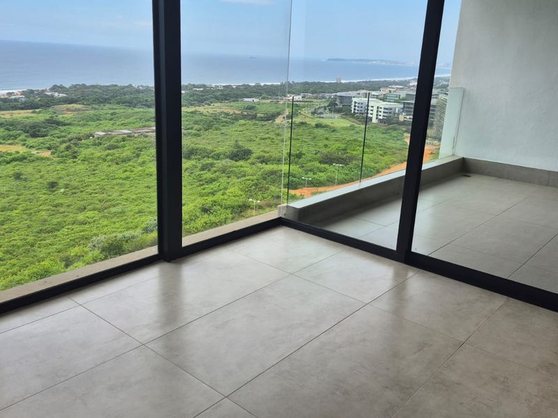 A modern, empty balcony featuring large floor-to-ceiling glass sliding doors, tiled flooring, and a glass balustrade with expansive coastal and landscape views.