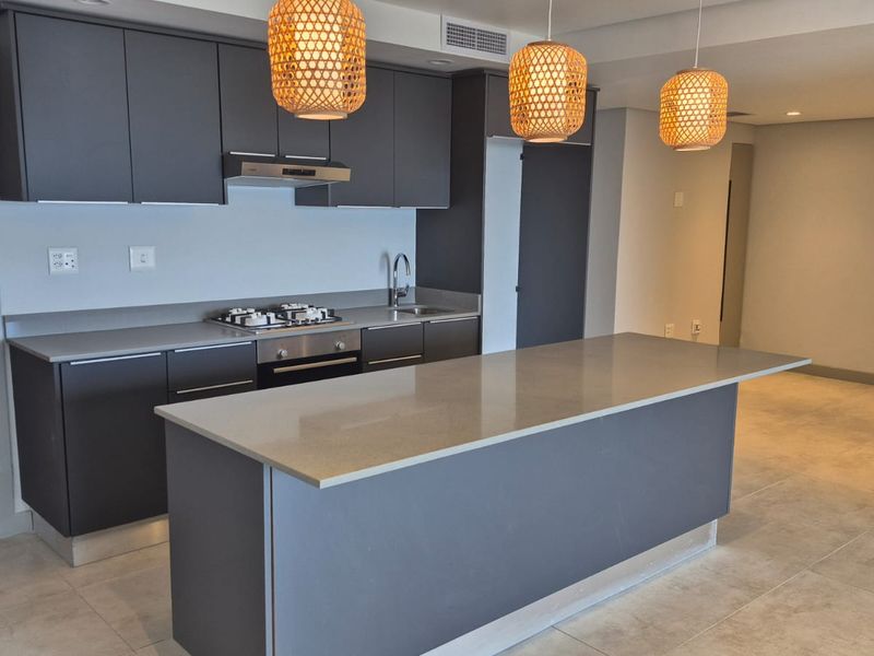 Modern open-plan kitchen featuring dark cabinetry, stone countertops, a central island, and integrated appliances, seamlessly flowing into the adjacent living area.