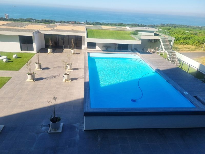 An elevated view of a modern apartment complex pool area featuring a tiled deck, glass balustrades, and a well-maintained swimming pool with ocean views.