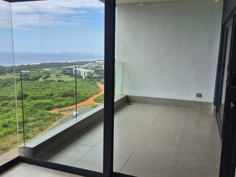 A modern, spacious balcony featuring large-format floor tiles, a frameless glass balustrade, and expansive views of the surrounding landscape and coastline.