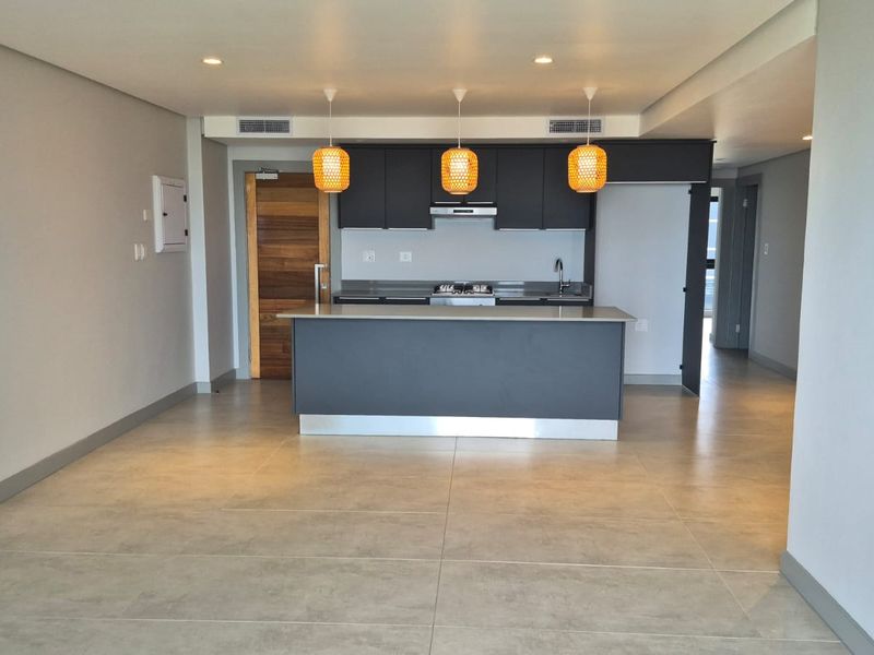 Modern open-plan living and kitchen area featuring sleek dark cabinetry, a central island, tiled flooring, and recessed lighting in a contemporary apartment setting.