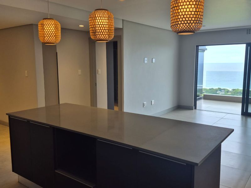 Spacious open-plan living area featuring a modern kitchen island, neutral tiled flooring, and large sliding doors opening to a balcony with ocean views.
