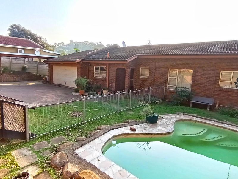 Rear view of a brick house featuring a swimming pool, paved driveway, attached garage, and metal perimeter fencing.