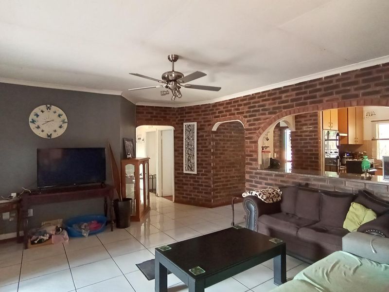 Open plan living area featuring tiled flooring, exposed brick walls, and a ceiling fan, with a pass-through window connecting to the kitchen.