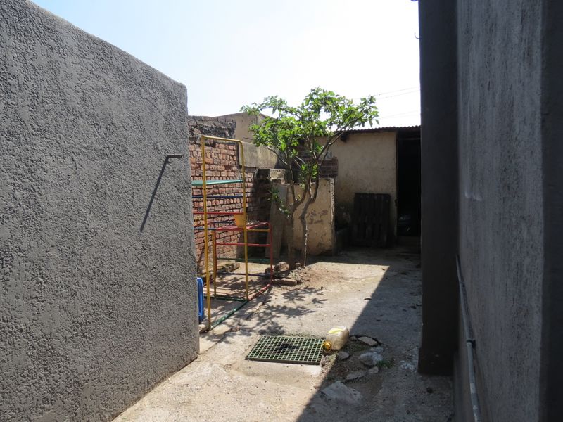 A narrow rear exterior area featuring a concrete pathway, brick boundary walls, and a drainage grate, requiring maintenance and landscaping improvements.