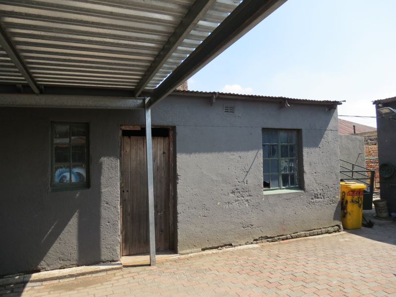 Rear exterior view of a dwelling featuring a corrugated metal roof, steel carport, and paved courtyard area with visible maintenance requirements.