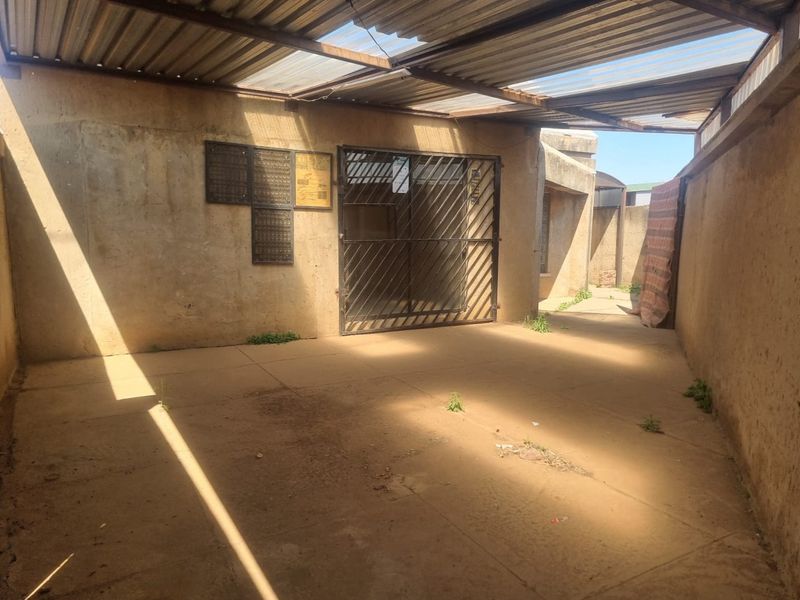 A covered outdoor patio area featuring concrete paving, a security gate, and corrugated roof sheeting, requiring maintenance and surface repairs.