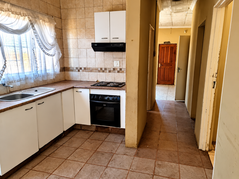 A functional kitchen featuring built-in cabinetry, a stainless steel sink, and an integrated oven with hob, leading into a tiled hallway.