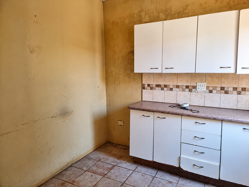A small kitchen area featuring white built-in cabinetry, a tiled backsplash, and tiled flooring, currently in need of cosmetic refurbishment and wall repairs.