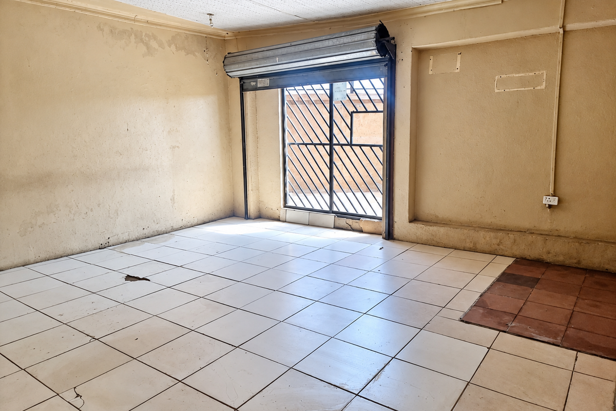 An empty, dated living area featuring tiled flooring, a security roller shutter, and a security gate, requiring significant cosmetic renovation and repair.