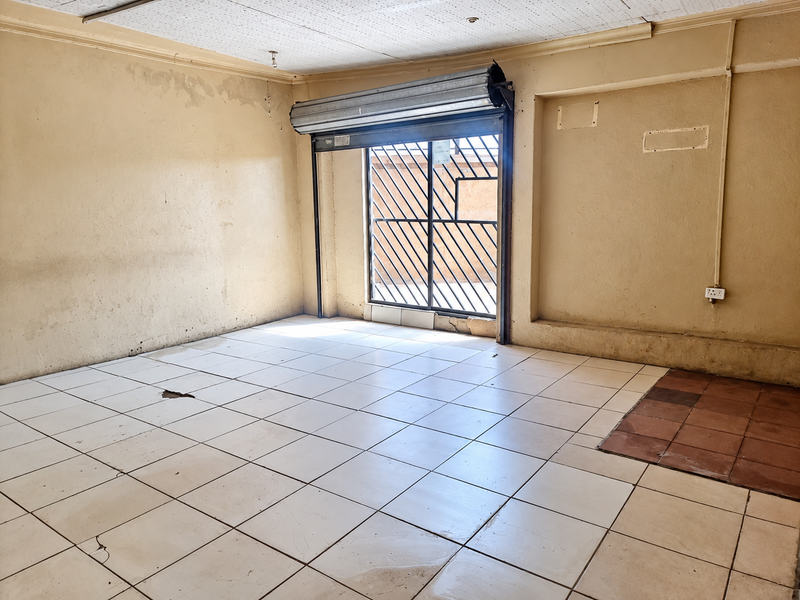 An empty, dated living area featuring tiled flooring, a security roller shutter, and a security gate, requiring significant cosmetic renovation and repair.