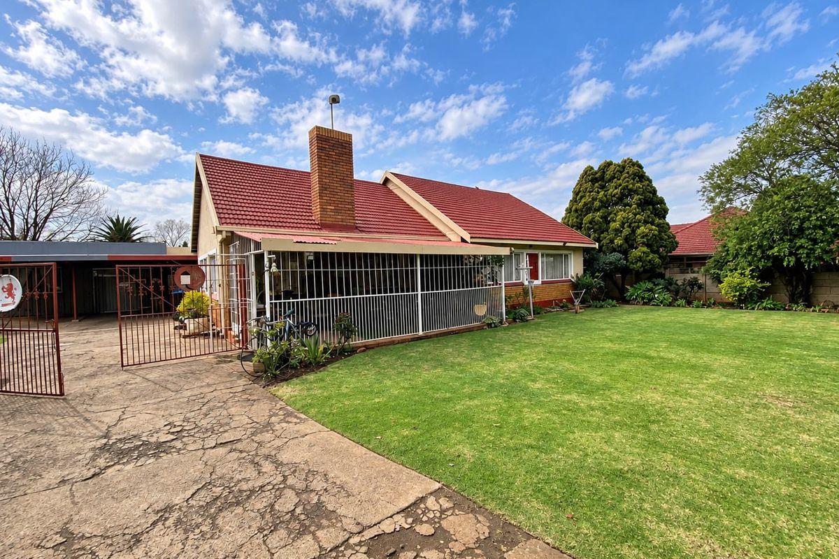 Rear view of the house featuring a tiled roof, brick chimney, covered patio with security bars, and a spacious lawn with a cracked concrete driveway.