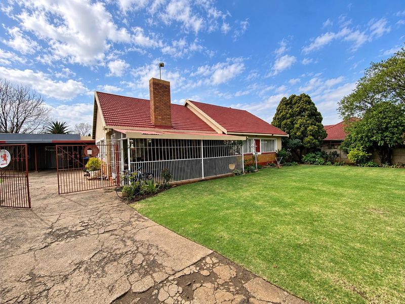 Rear view of the house featuring a tiled roof, brick chimney, covered patio with security bars, and a spacious lawn with a cracked concrete driveway.