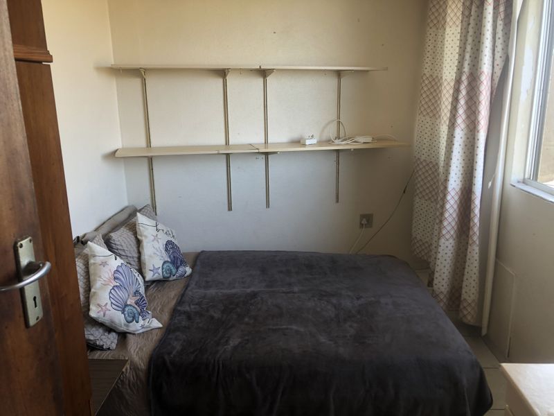 A compact bedroom featuring wall-mounted shelving and a window with curtains, requiring minor cosmetic updates to improve the overall finish.