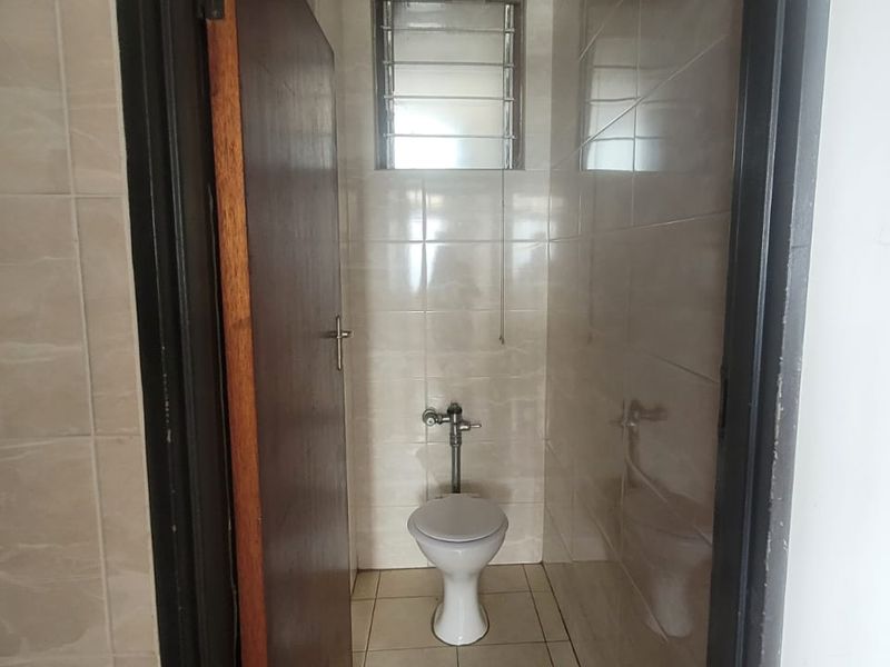 A compact, tiled bathroom featuring a standard toilet and a louver window for ventilation, adjacent to the kitchen area.