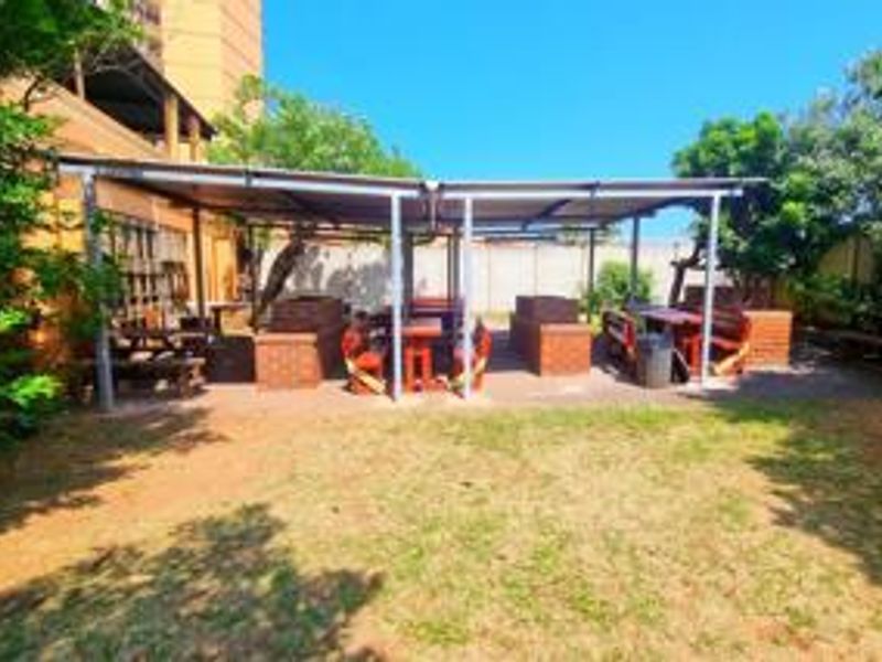 A communal outdoor entertainment area featuring a covered patio with built-in brick braai facilities and paved flooring, set within a garden space.