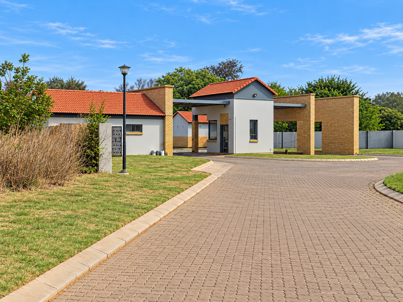 A well-maintained residential estate entrance featuring a paved driveway, security gatehouse, and brick boundary walls under a clear blue sky.