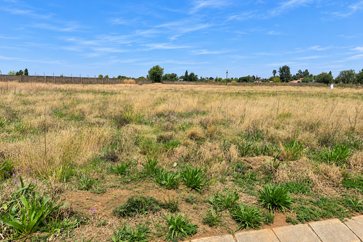 A vacant residential plot featuring natural grass coverage, a perimeter boundary wall, and a paved verge, offering a clear space for future development.
