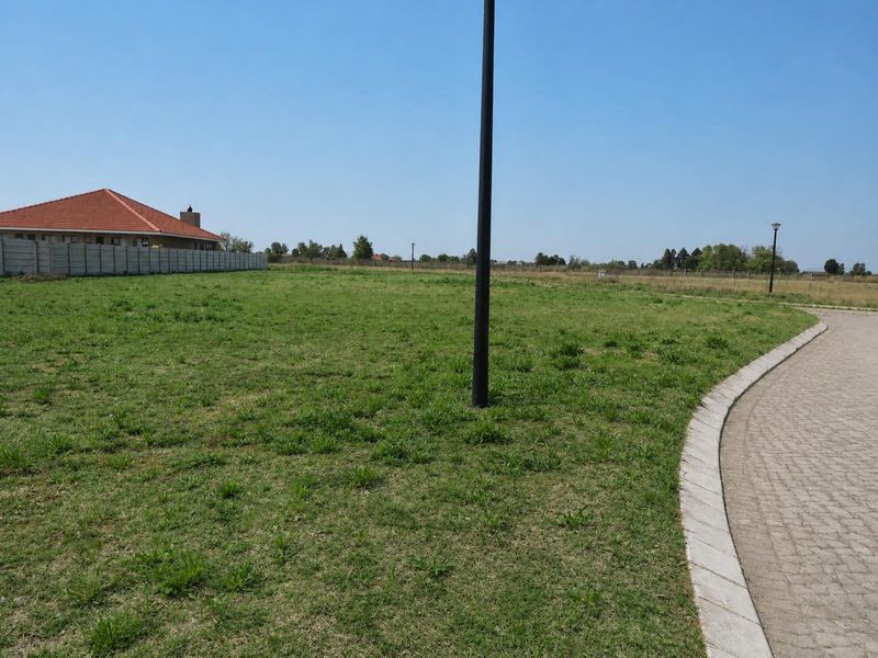 A vacant residential plot featuring a paved driveway edge, boundary wall, and established grass, offering a clear space for future development.