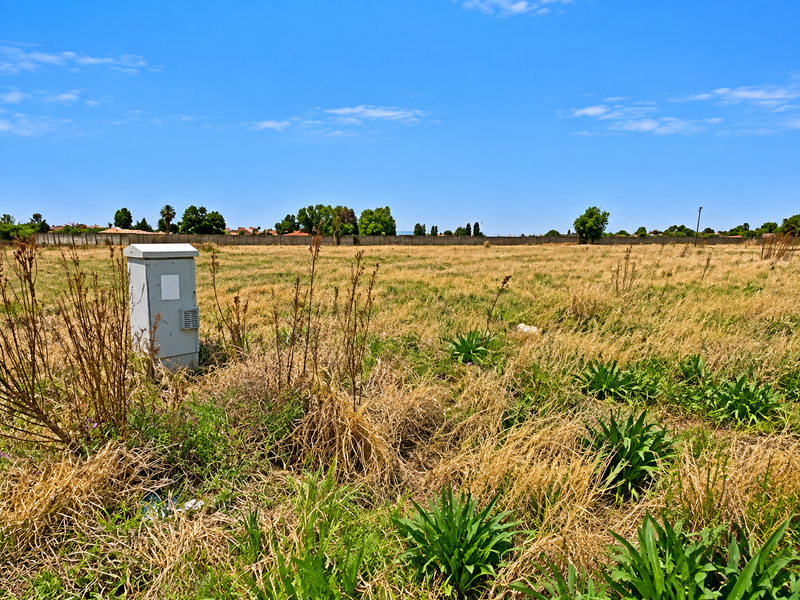 A vacant residential plot featuring dry grass, a perimeter boundary wall, and an installed electrical utility box.