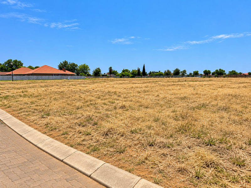 A vacant residential plot featuring dry grass, a paved curb, and surrounding boundary walls in a developed neighborhood.